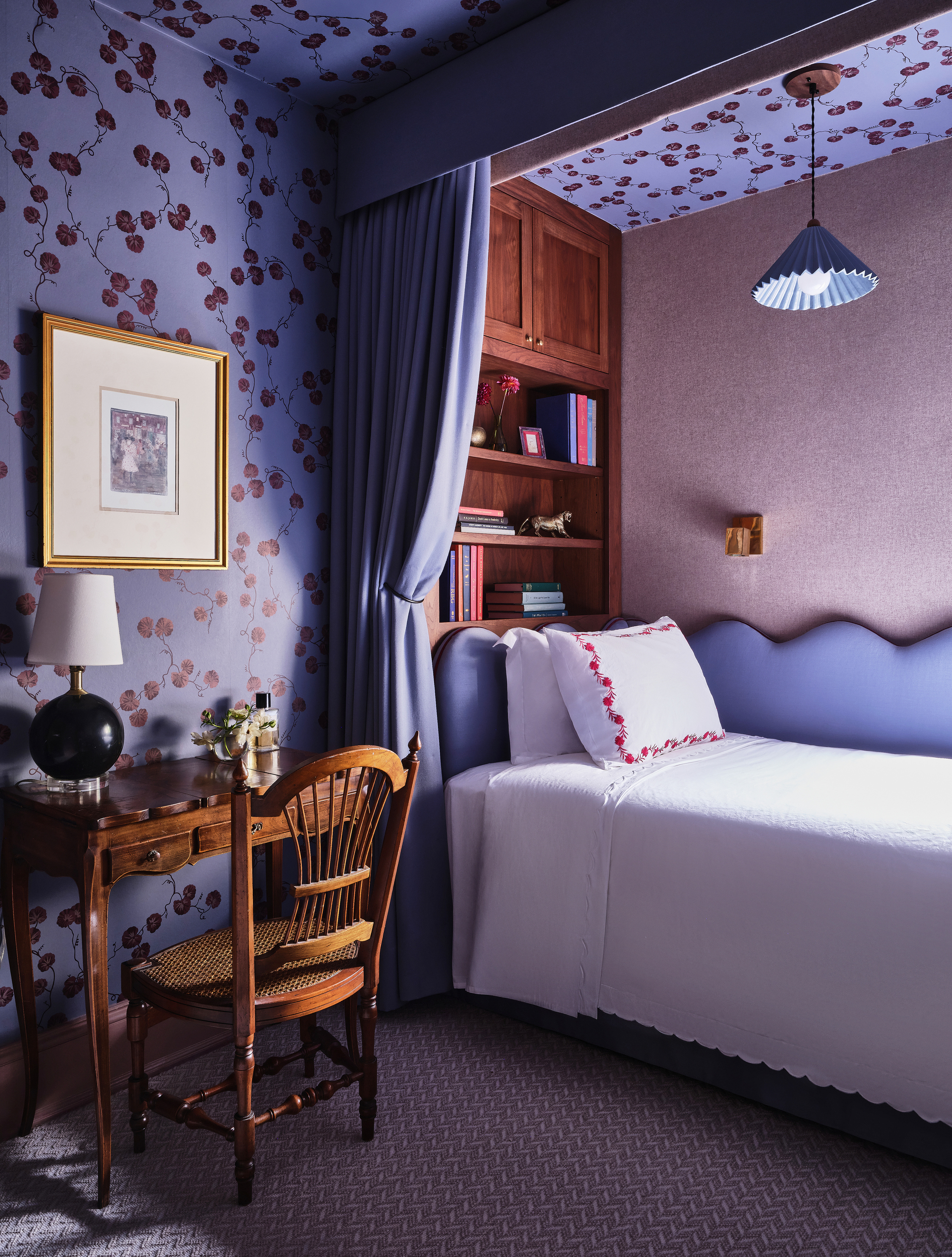 purple bedroom with alcove bed with curtain, scalloped headboard, built-in book shelves, an antique timber desk and chair with table lamp and artwork on the wall above