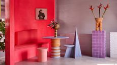 Image of a light, lavender room with a hot pink bench seat. There is a periwinkle blue round table and geometric chair, a light pink stool, and a purple tile stand with an orange vase on it also in the room.