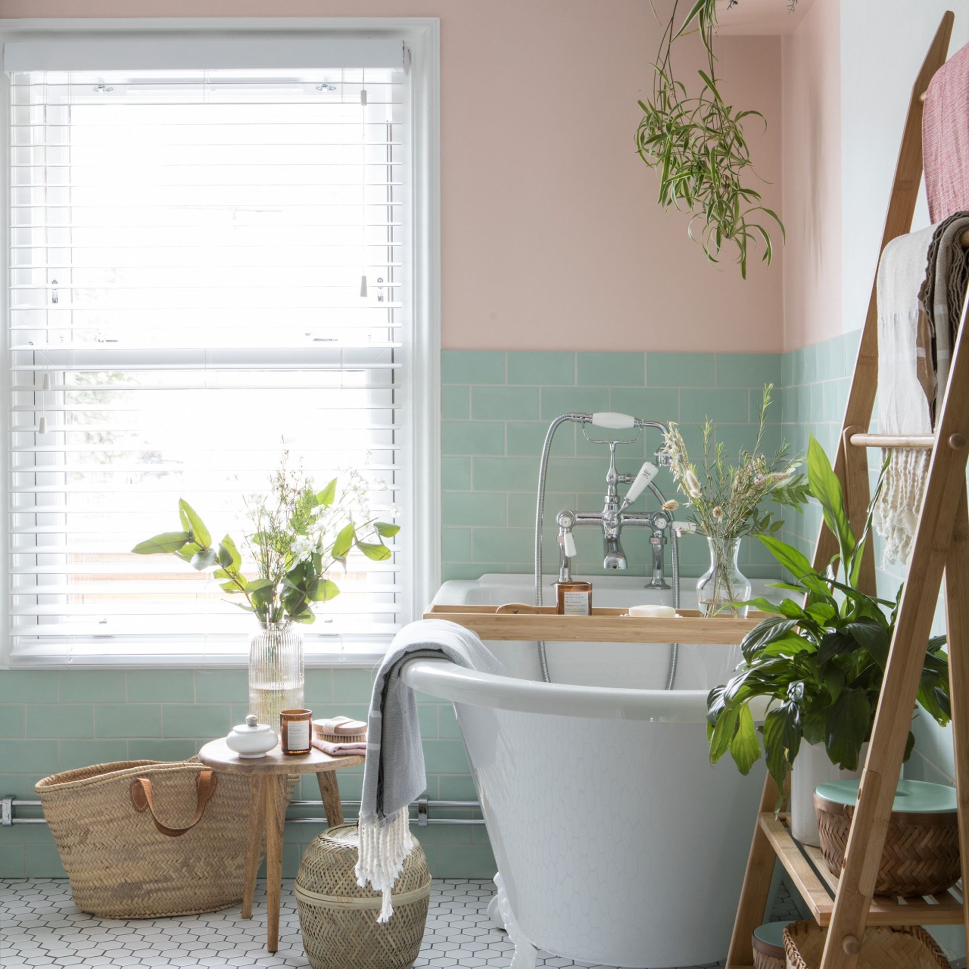 Pink and blue tiled bathroom with a white bathtub, decorative storage baskets and houseplants with a large window and blinds
