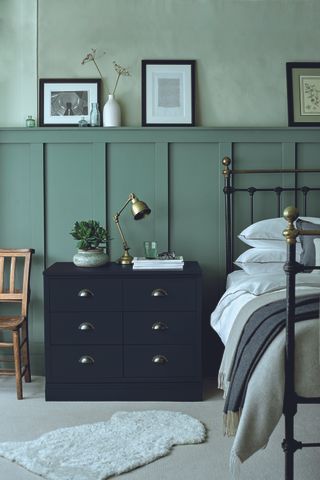 two tone green bedroom with black iron bed, black chest of drawers, painted paneling, artwork on shelf, white bedding, chair, brass desk lamp