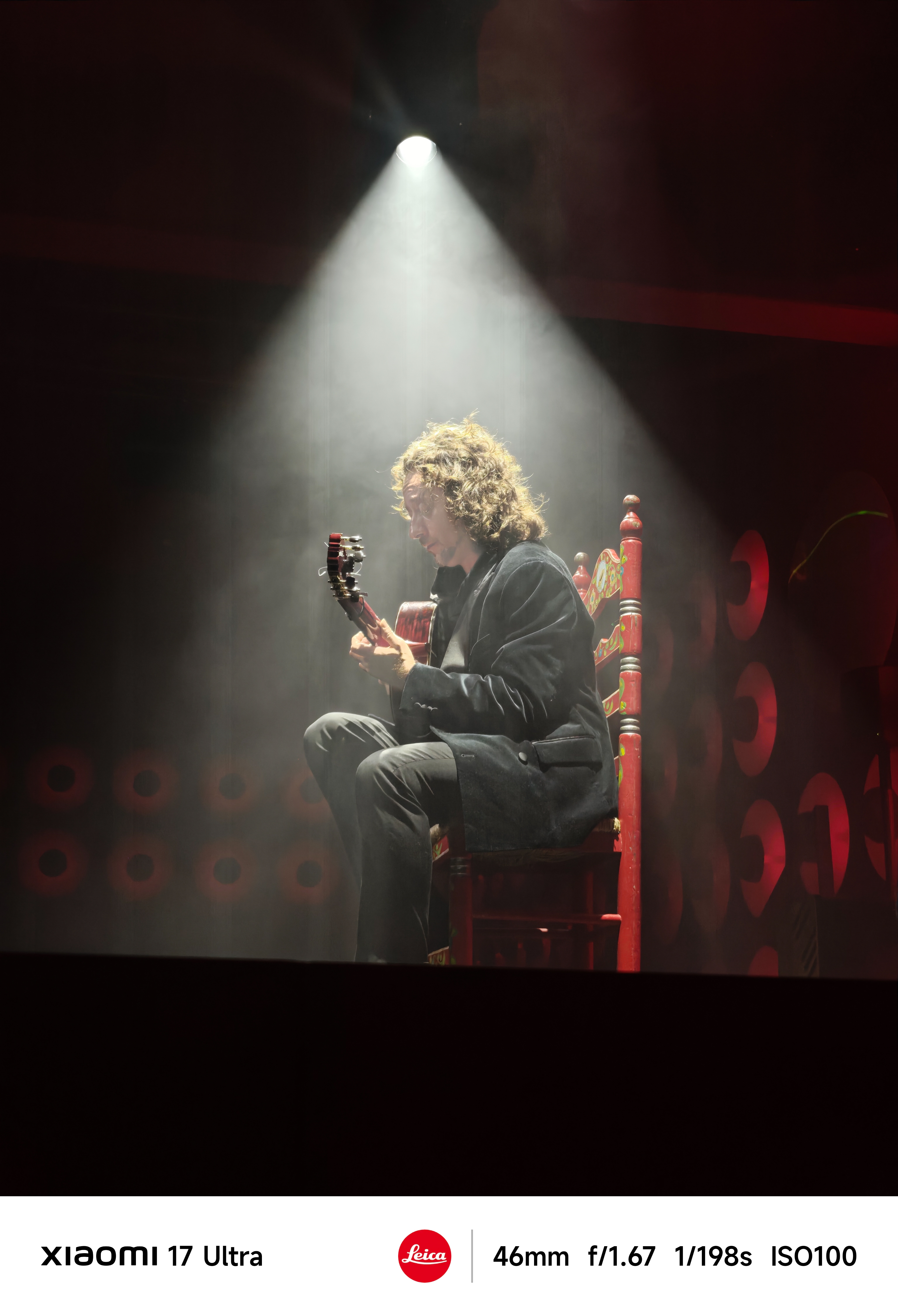 Flamenco guitarist seated on a red chair under a dramatic spotlight on stage.