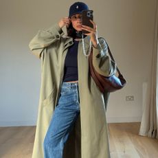 Influencer takes a mirror selfie wearing jeans, heels high vamp shoes and a khaki trench coat.