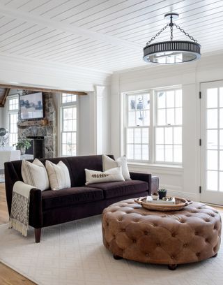 white living room with shiplap walls, wooden floor, brown couch, brown leather round footstool, cream rug