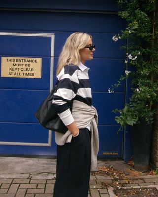 British fashion influencer Lucy Williams poses on a sidewalk wearing sunglasses, a black and white stripe rugby polo shirt, black leather bag, tan sweater tied around the waist, and black pants