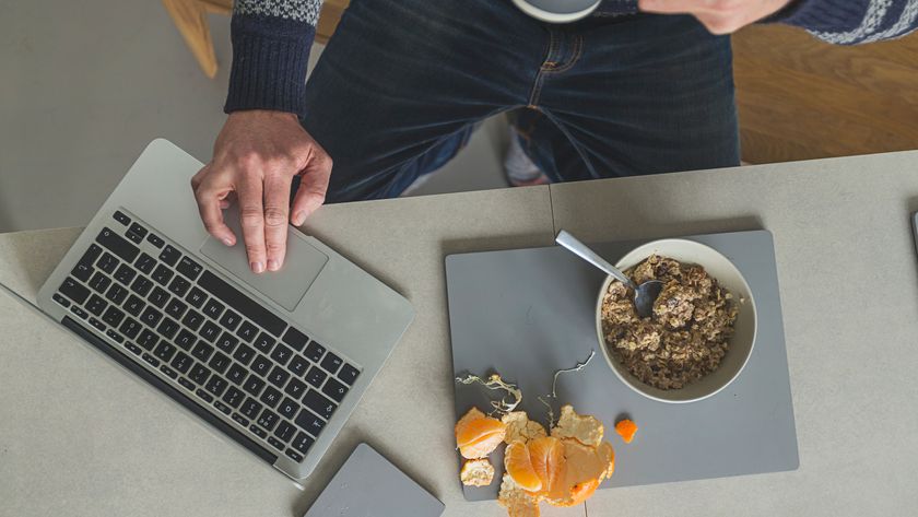 A person sat at their home desk using a laptop, with a mug of coffee in one hand and a cereal bowl and orange peel on their desk, shot from above.