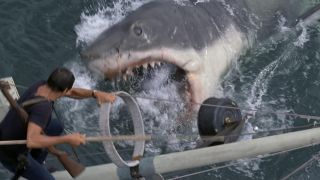 Still from the movie Jaws. Here we see a giant great white shark breaching the waves to attack a boat. A man with a shotgun on his back is scrambling up the boat's mast to get to safety.