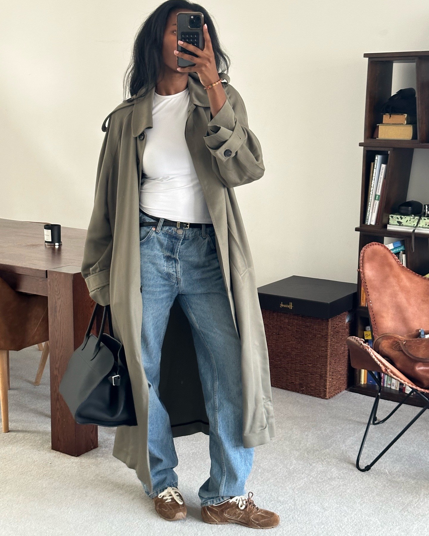 Fashion influencer Marilyn Nwawulor-Kazemaks in London taking a mirror selfie wearing a chic fall outfit with a relaxef trench, white tee, black tote, suede sneakers, baggy jeans and a skinny belt.