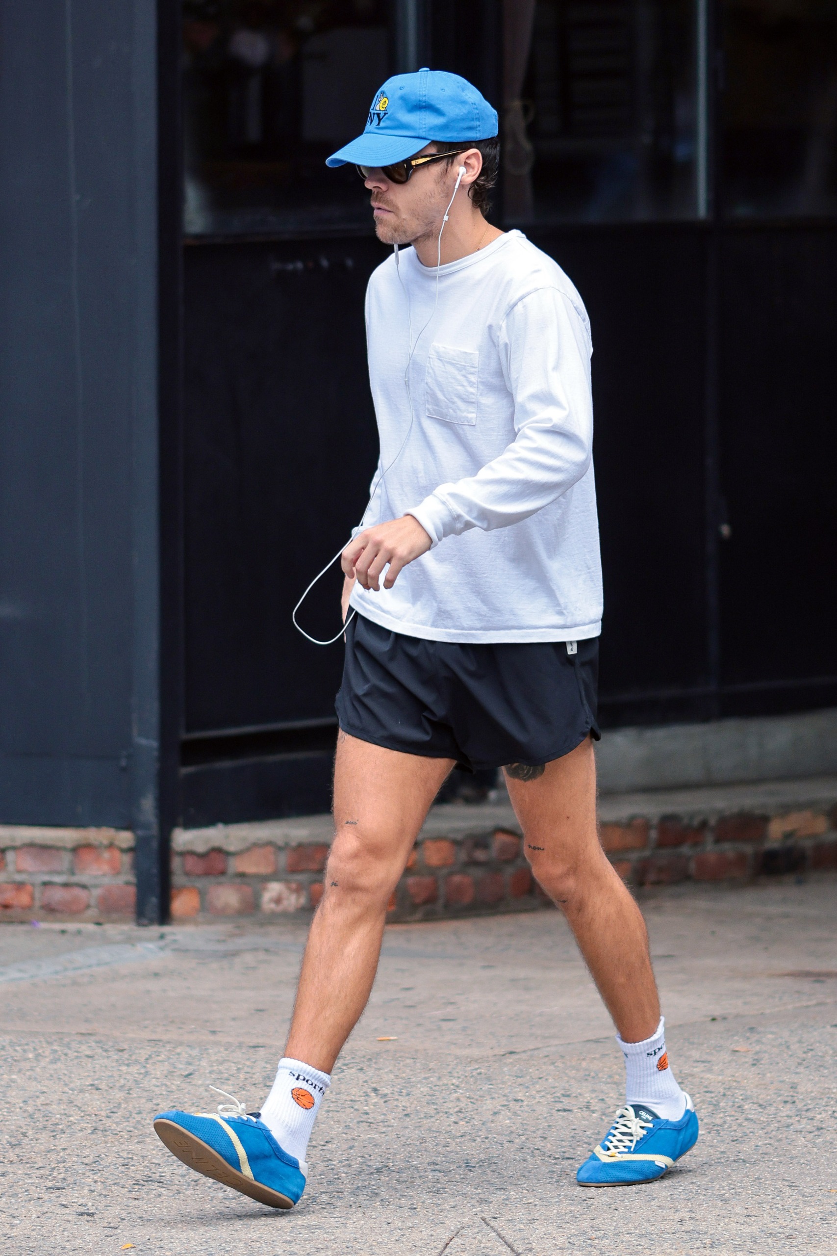An image of Harry Styles wearing a long-sleeved shirt with track shorts and Celine Flat Sneakers whilst in New York.