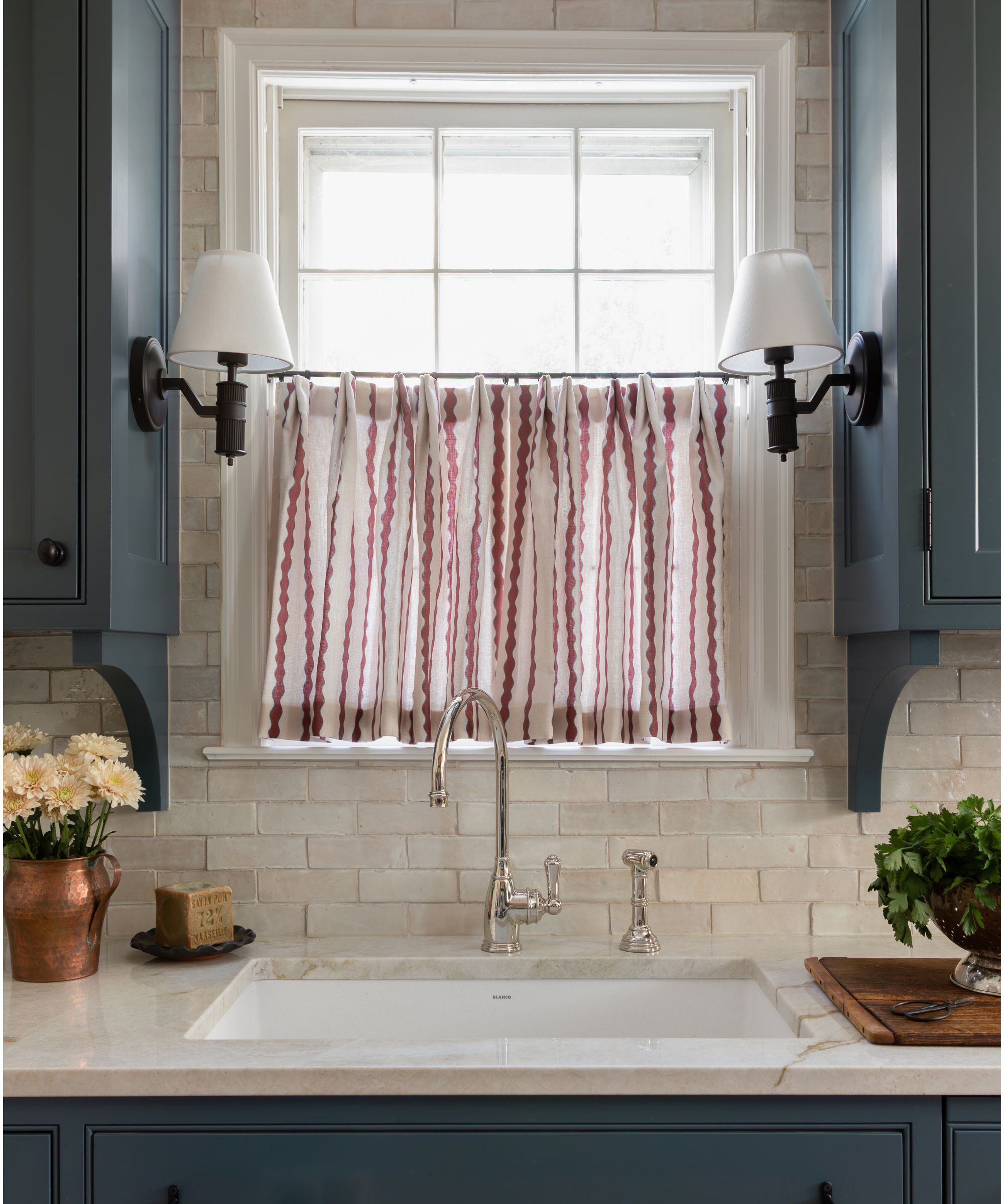 kitchen window dressed with a white and red striped cafe curtain, blue painted cabinets either side of the window with small black and white sconces
