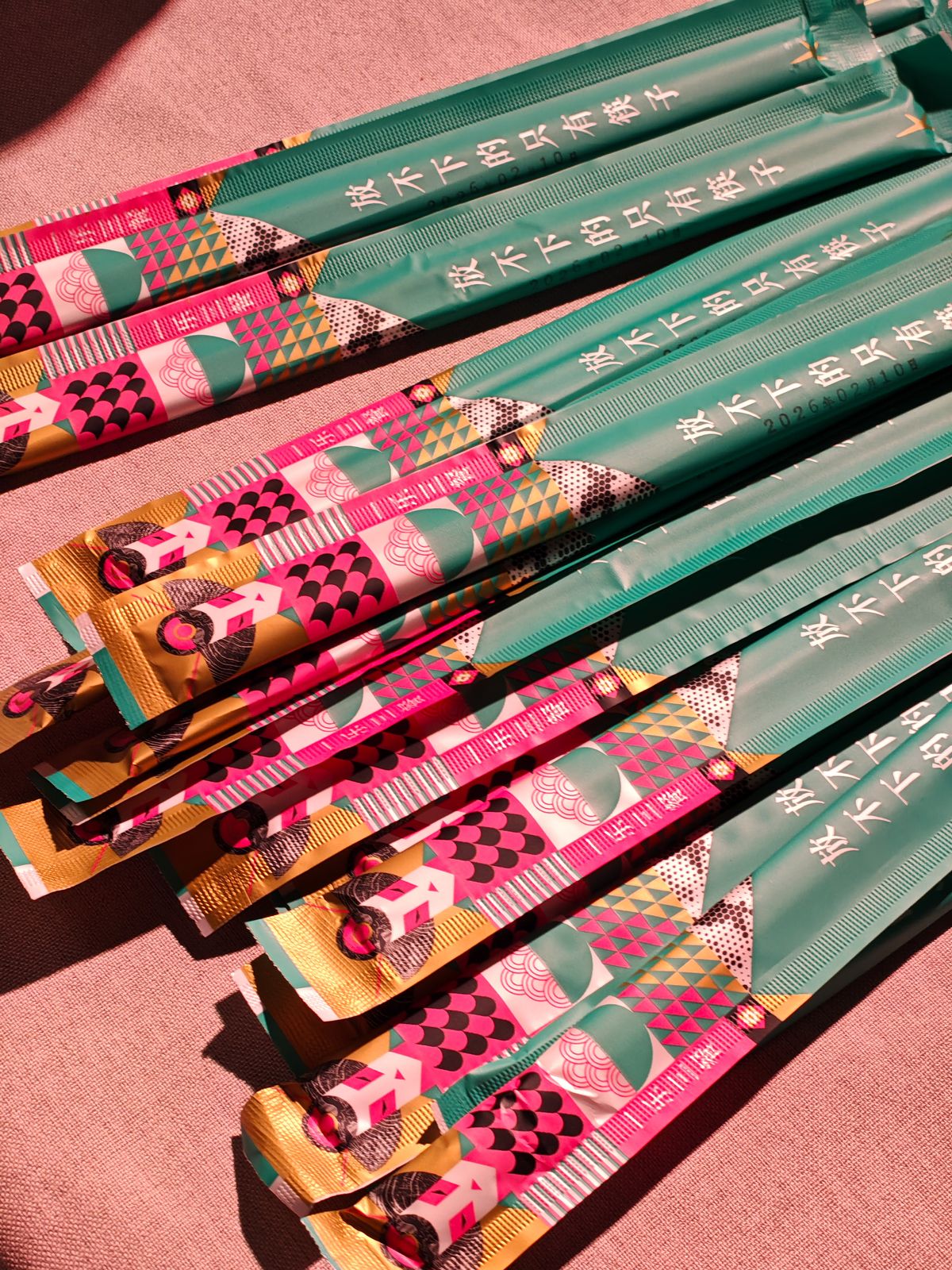 A close-up, high-angle shot of several pairs of disposable chopsticks in decorative paper sleeves. The sleeves feature a vibrant, colorful geometric pattern in shades of teal, pink, gold, and black. Chinese text is printed on the teal section of the sleeves. The items are laid out on a textured pink surface.