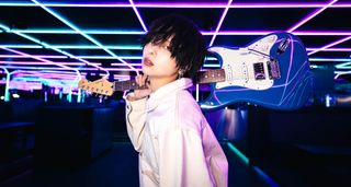Otoha holds a blue Fender Strat in a staged setting with neon pink and blue lights overhead.