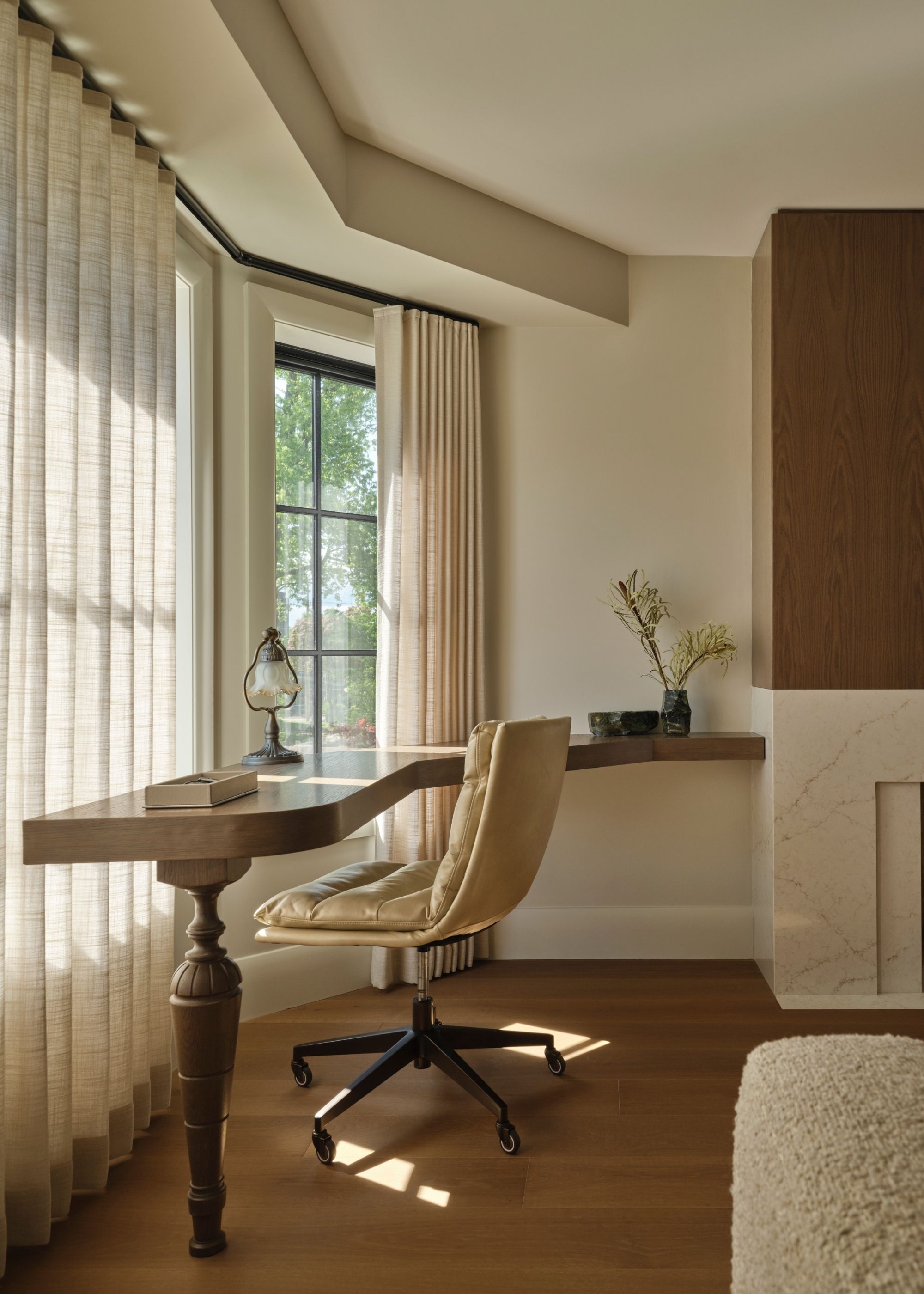 A minimalist home office with a floating desk, a leather swivel chair, beige curtains, a vase of stems, and a table lamp