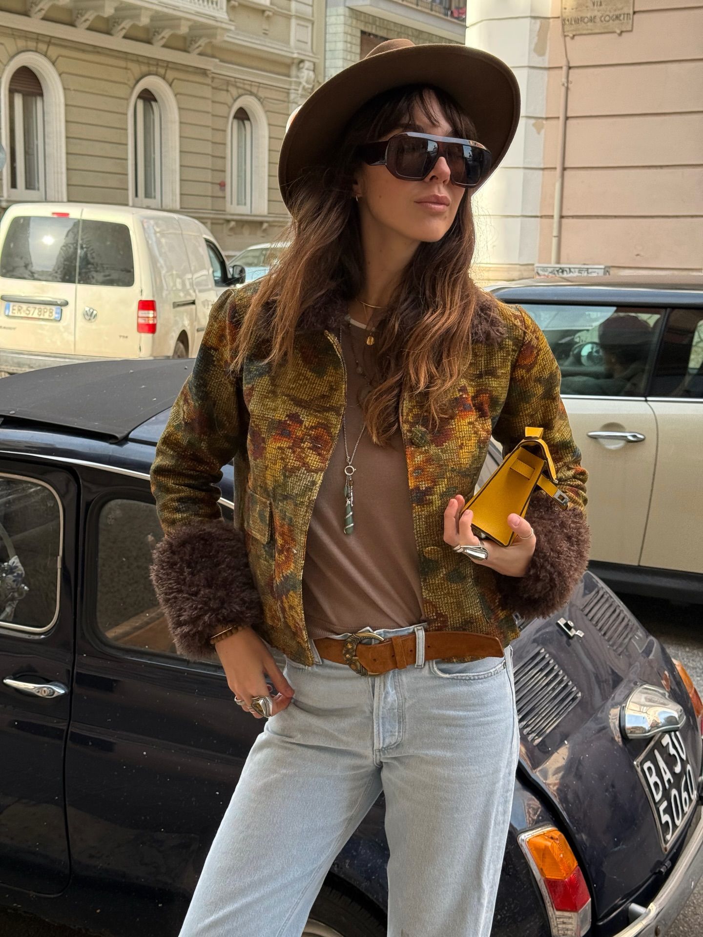 An image of a French influencer wearing a brocade jacket with faux fur cuffs over a brown shirt with light jeans and a felt hat holding a yellow handbag