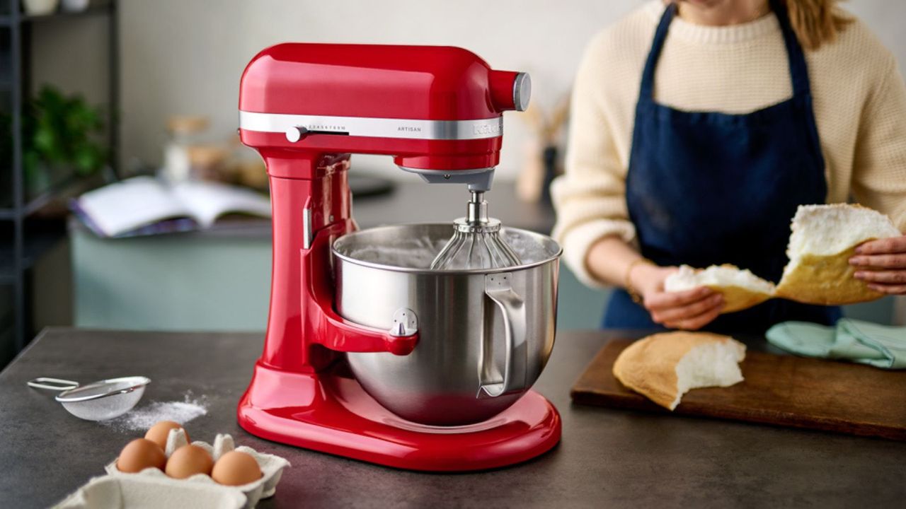 A carton of eggs beside a red KitchenAid stand mixer on a kitchen island; in the background, a woman tearing into a loaf of broad.