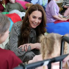 Kate Middleton wearing a black plaid dress with a tie neck kneeling on the floor talking to a little girl with families sitting behind her on the floor at a hospice