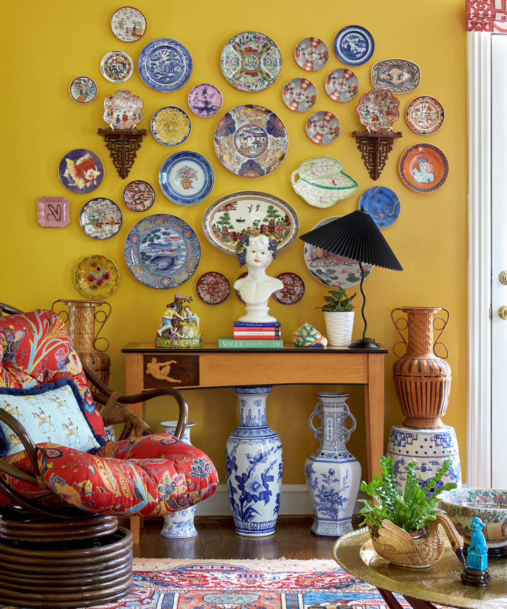 yellow living room with a gallery wall of china plates, a console table with objects and blue and white chinoiserie vases stood below with a red and dark wood armchair styled nearby
