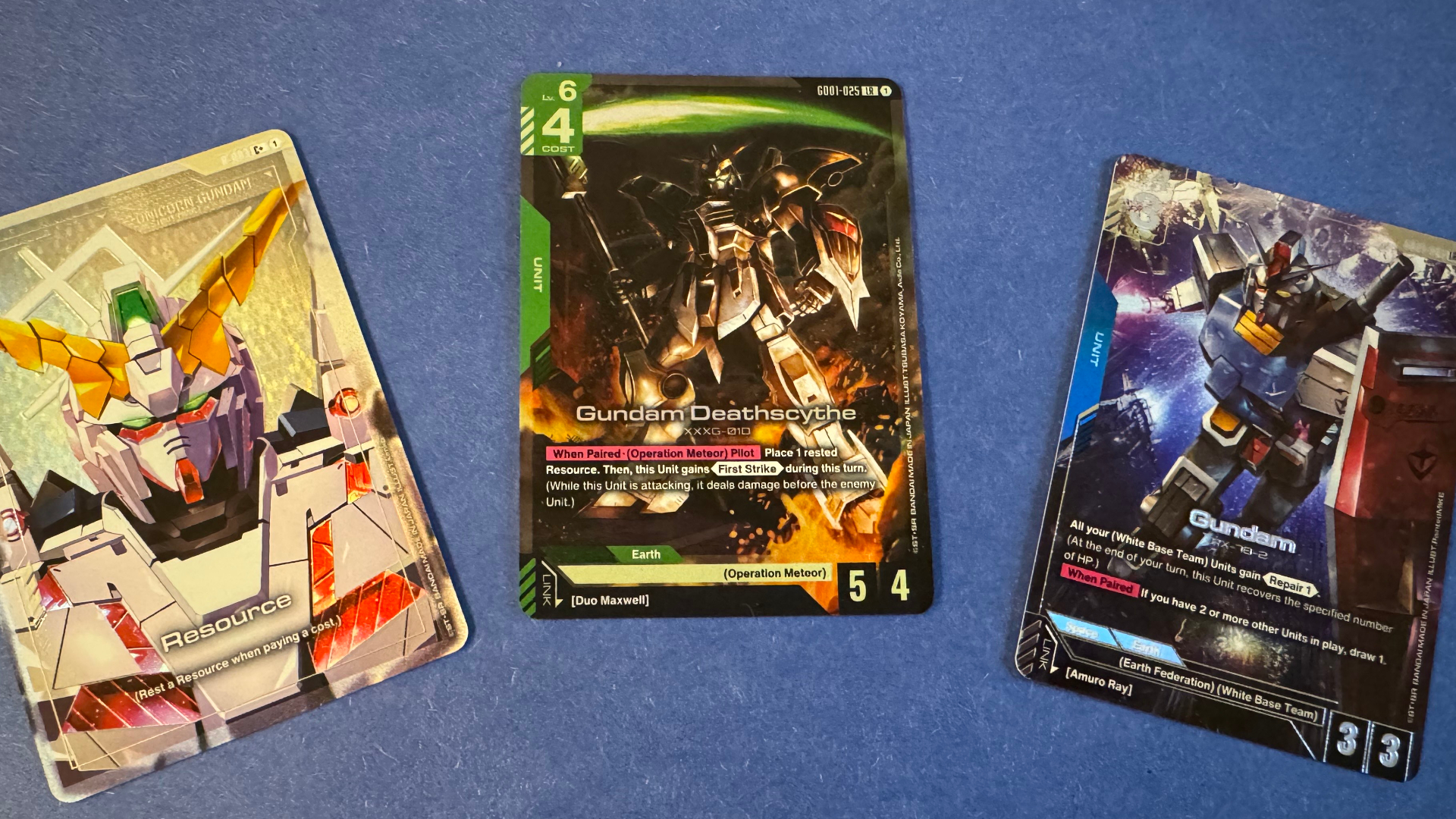 Three cards featuring Gundam mechs in various poses lie on a blue, plush surface