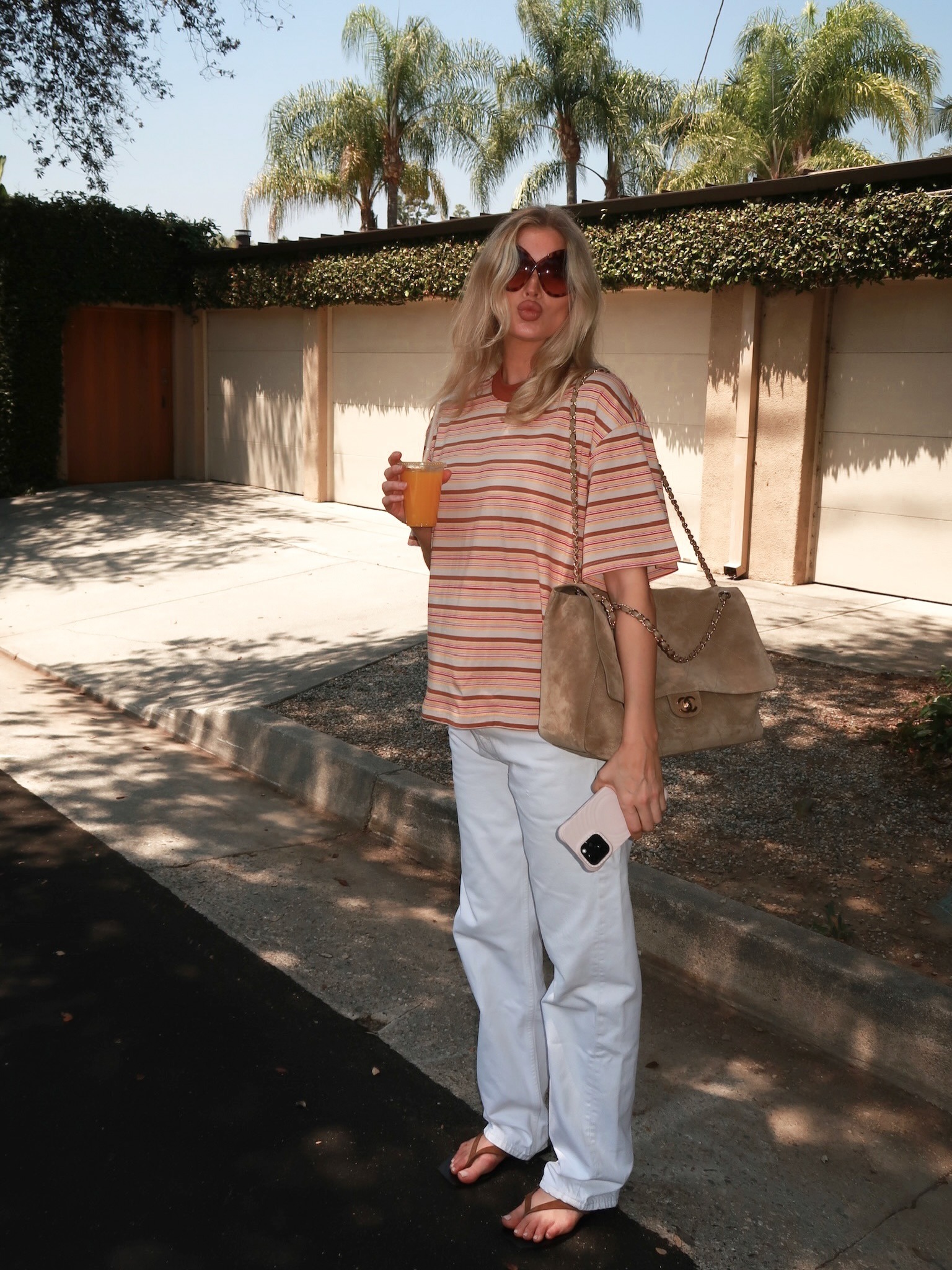 Elsa Hosk holds an orange juice wearing a striped t-shirt with white jeans and brown flip-flops.