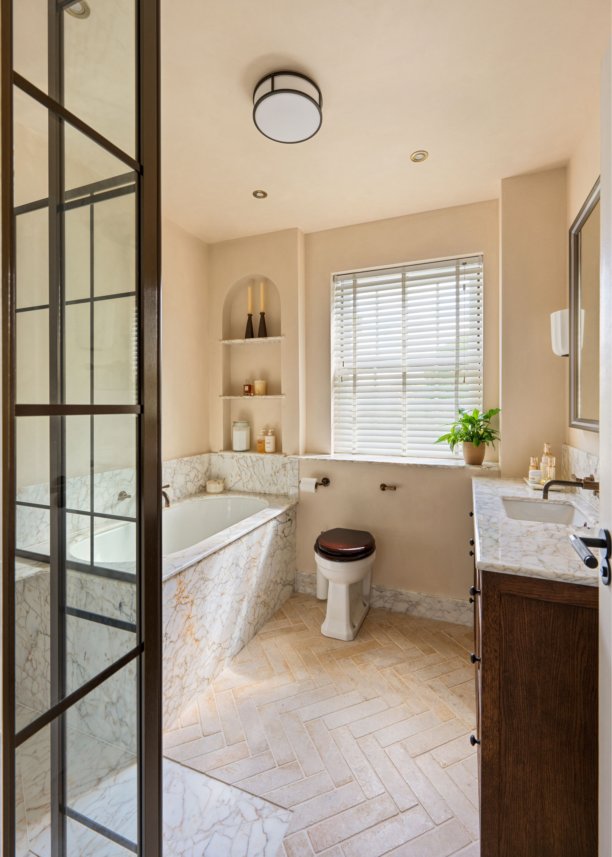 A bathroom with herringbone tiled flooring, an arched tiled shelving nook, a single vanity and a tub