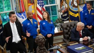 A man in a suit holds his hands apart, as if showing the long size of a subway sandwich. To the right, three people in blue flight jackets stand behind an old man at a desk.