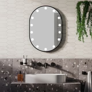 black framed oval mirror with LED circular lights around edge on white tiled wall above grey terrazzo effect tiles, white oval sink and wall mounted black taps