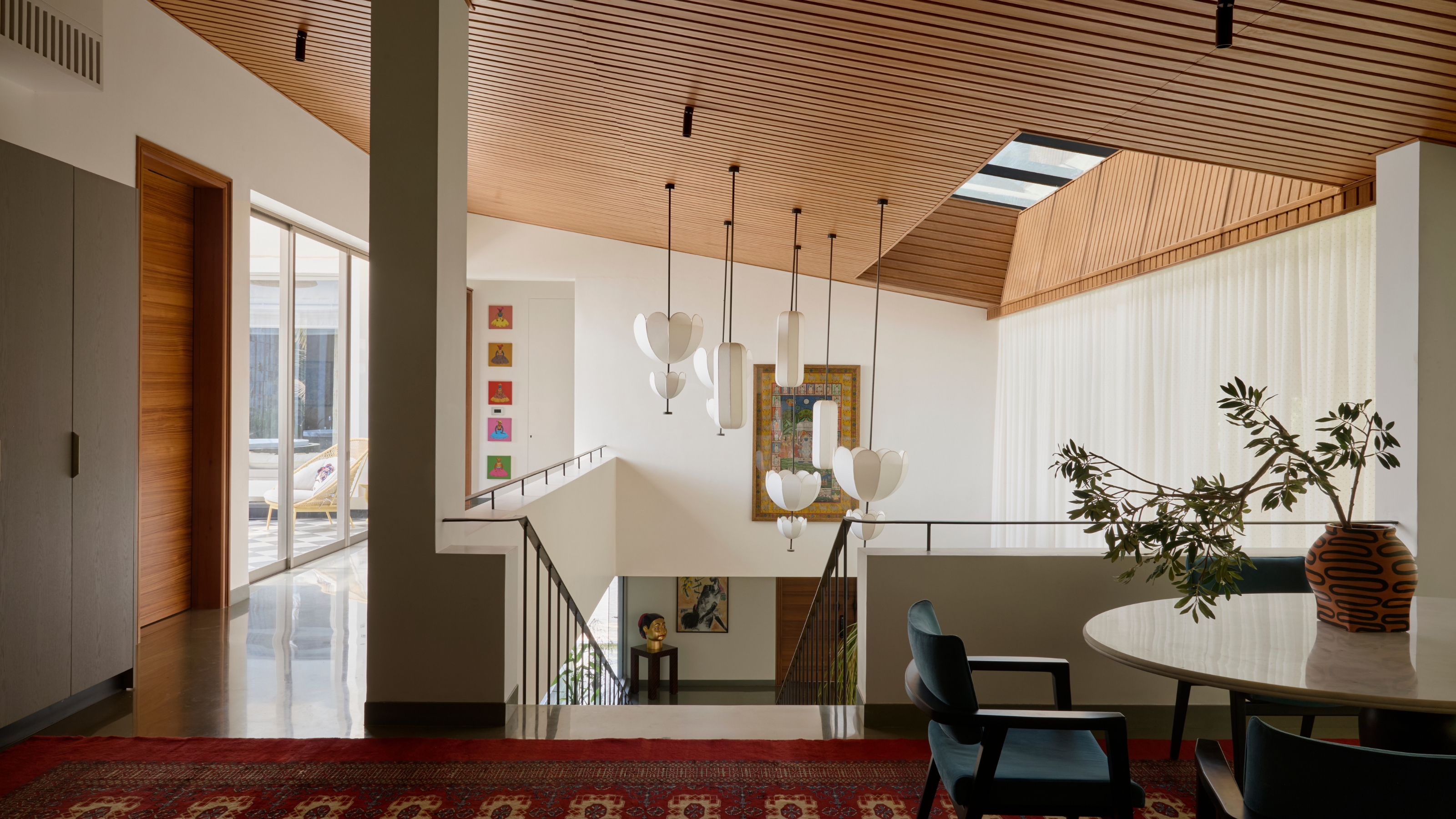 An upstairs area with an open concept staircase and skylight. The ceiling is wooden with a cluster of lantern pendant lights hanging by the staircase. There is art hung on the walls on both the upper and lower levels.