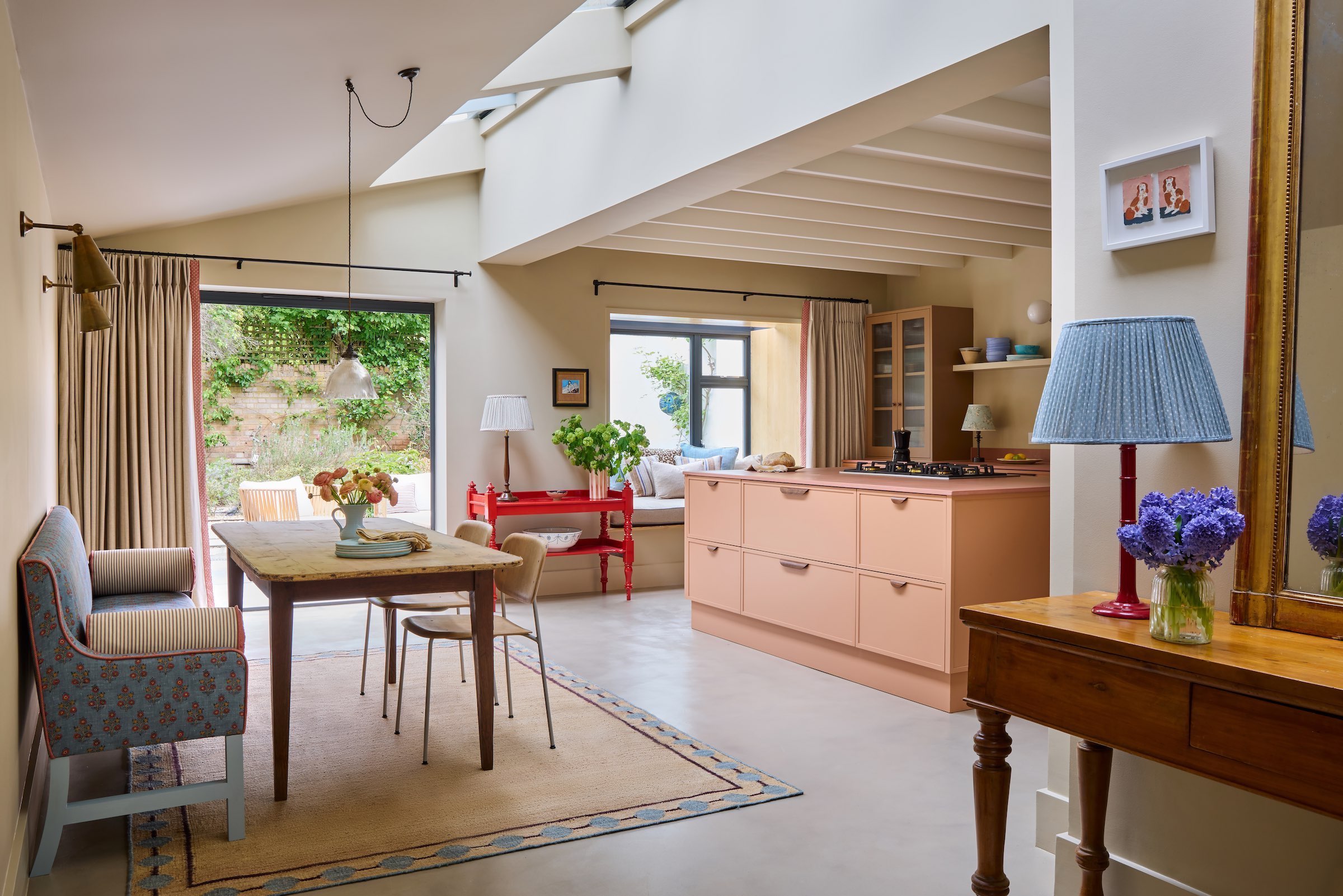 An open plan kitchen with a pink kitchen island that has extra storage. Opposite the island is a kitchen dining table with chairs.