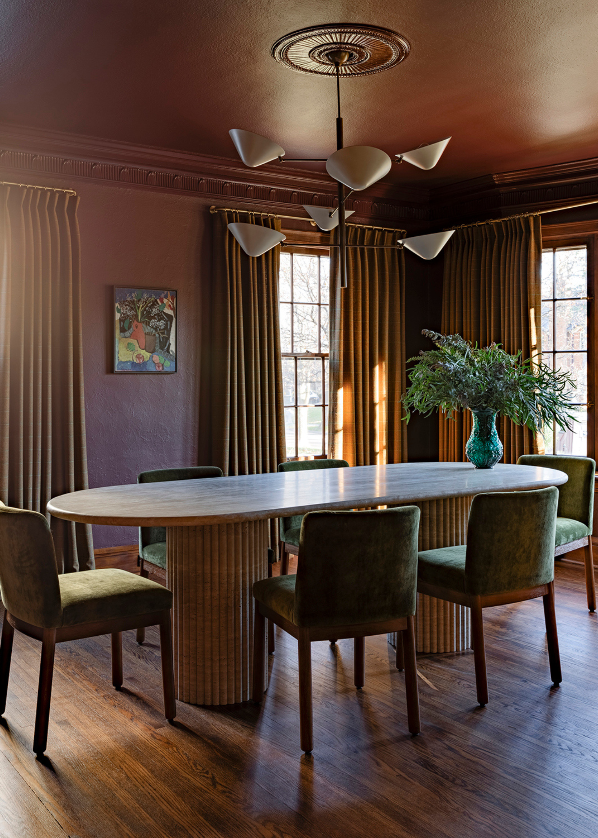 An oxblood-drenched dining room with ocher curtains, wooden flooring, a large oval dining table with two cylindrical ribbed legs, green velvet dining chairs, and a green vase of foliage atop the table