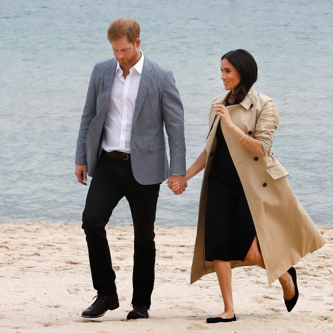 Meghan Markle walking on a beach holding hands with Prince Harry wearing a trench coat and black dress