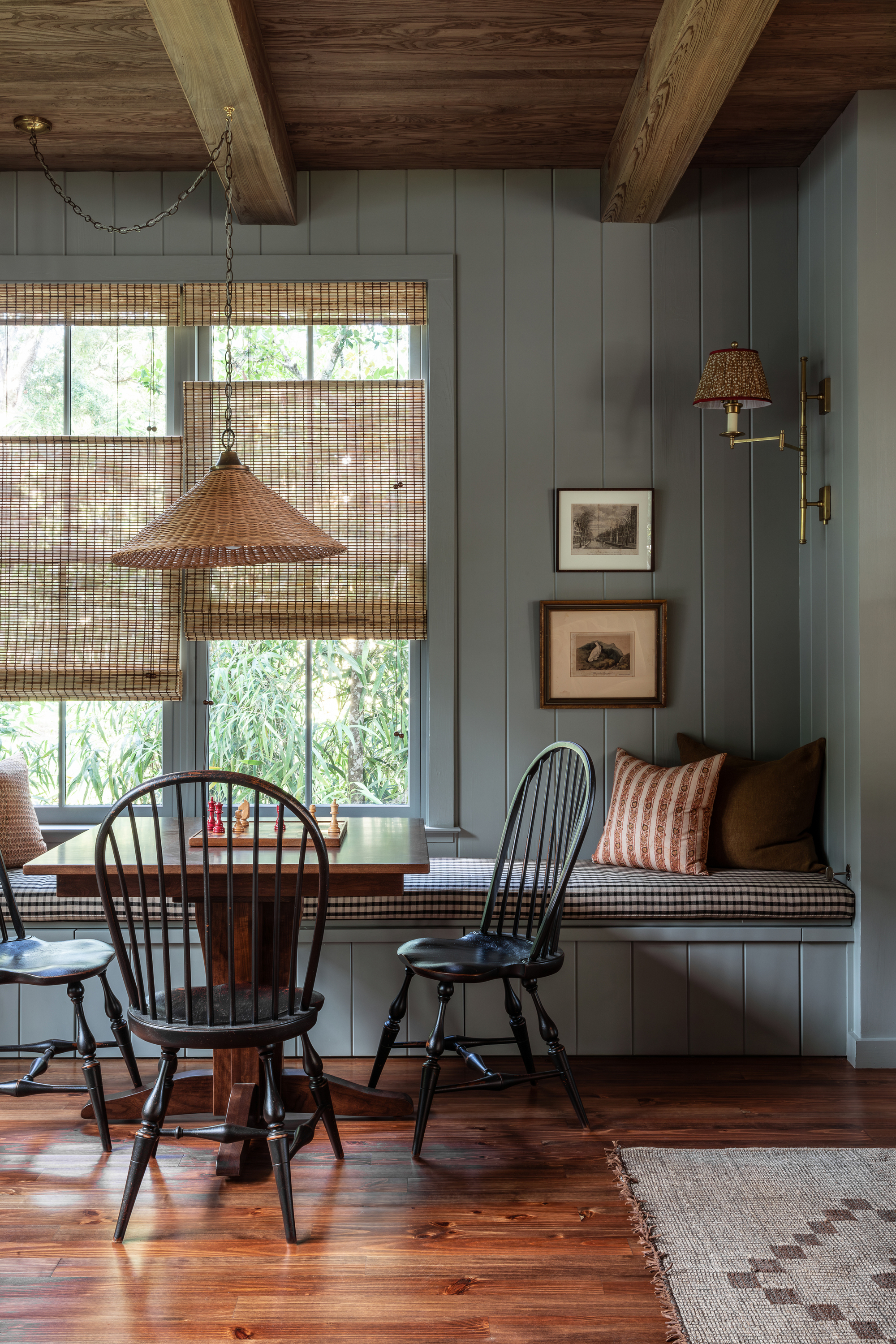 A mid-toned blue sitting area with a game table, tree spindle back chairs, and a built-in bench seat with a brown and white gingham cushion. There are rattan blinds on the window.