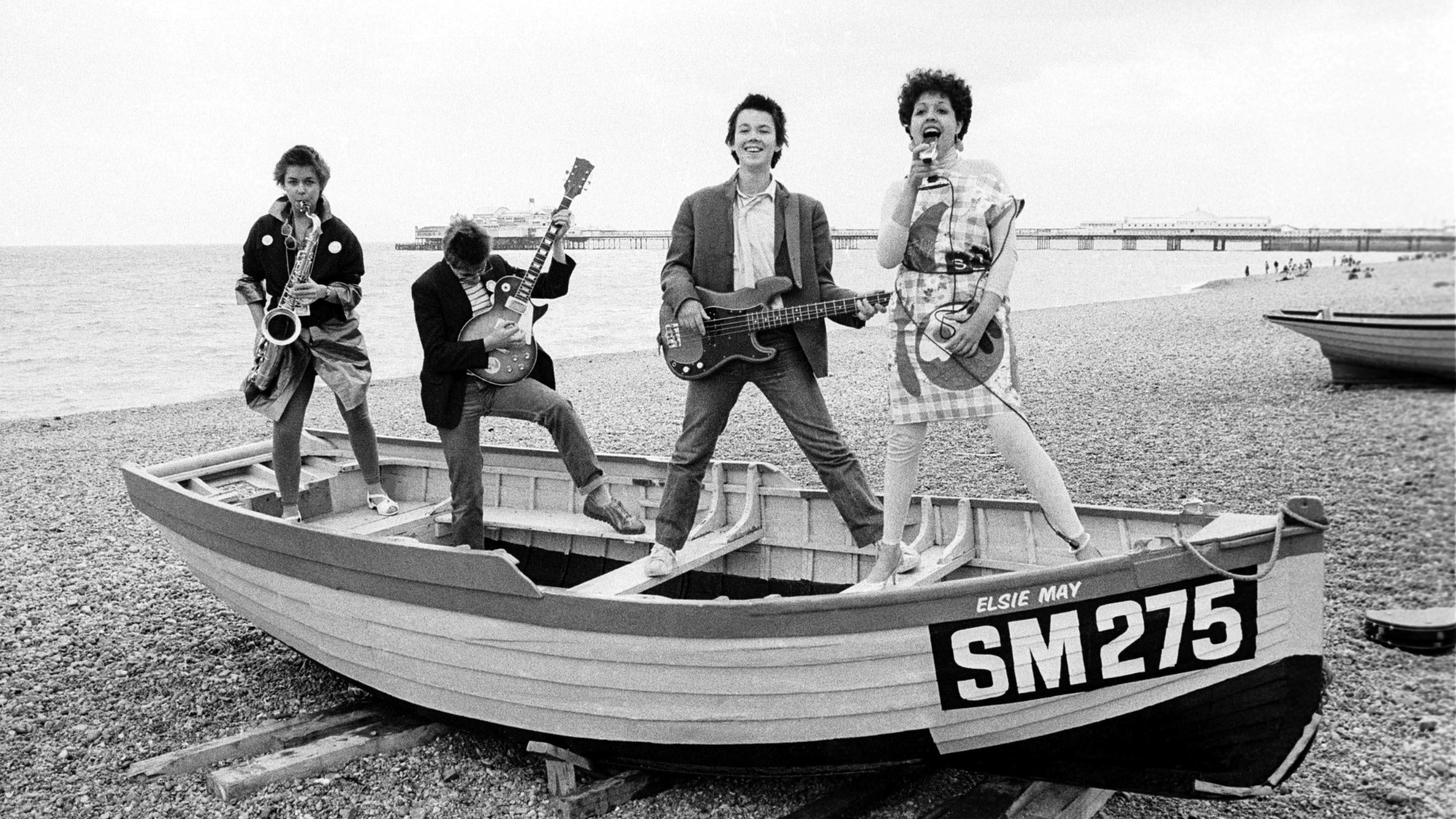 a black and white photo of X RAY SPEX on a small fishing boat on the shore at brighton beach in 1997. From L-R it shows Lora Logic, Jak Airport, Paul Dean, and Poly Styrene