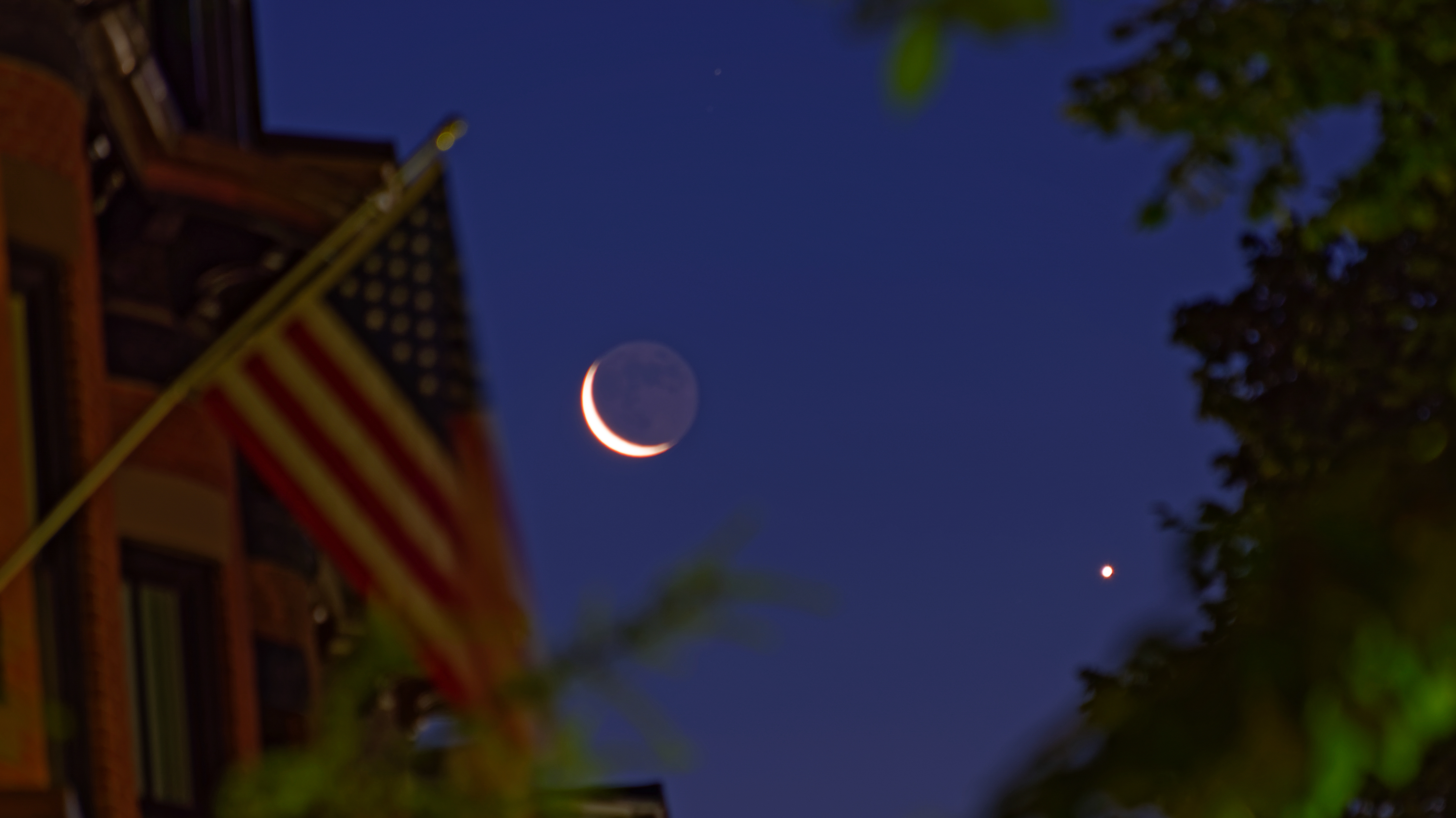 The rising moon in conjunction with Venus is framed on the left by brick townhouses with wooden dormers and an unfurled American flag in Boston's historic South End. Leafy treetops complete the framing on the right. The photo was taken at the start of nautical twilight.