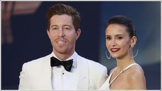 Shaun White wearing a white suit, smiling with Nina Dobrev - who is wearing a white satin gown, as they attend the "Top Gun: Maverick" Royal Film Performance at Leicester Square on May 19, 2022 in London, England.