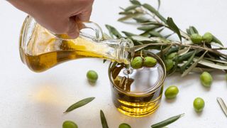 Woman's hand pouring olive oil from glass jug into glass, surrounded by olive branch