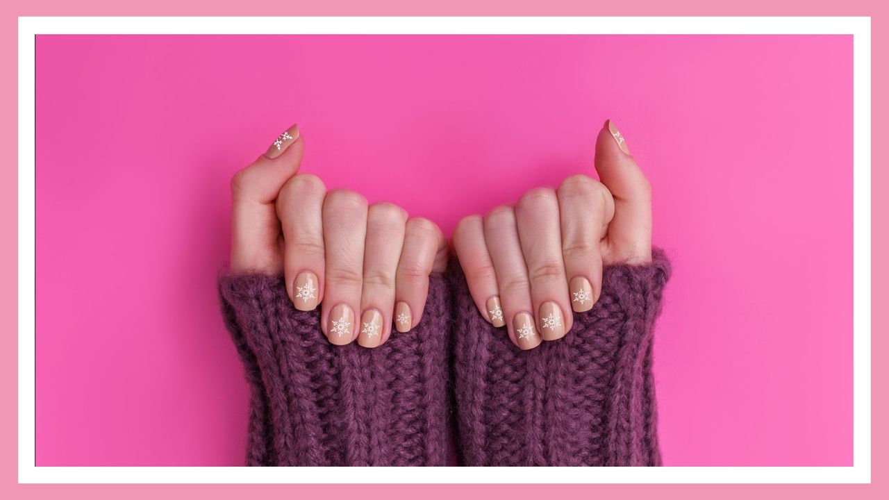 woman&#039;s hands showing off Christmas nails—a snowflake manicure on pink nails, against a pink background