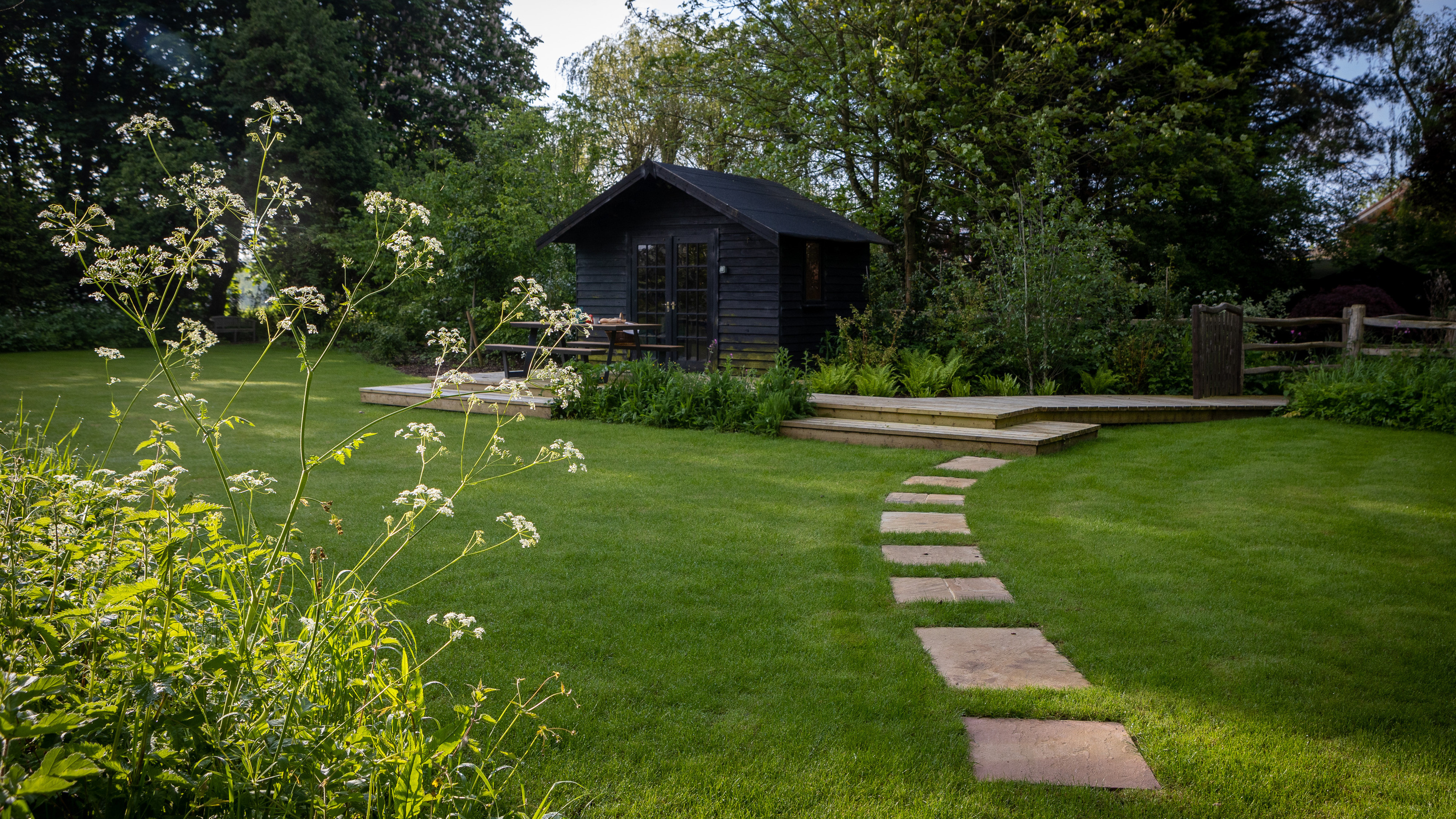 A garden with a large lawn, a paving stone path, and a garden shed painted black on a deck platform 