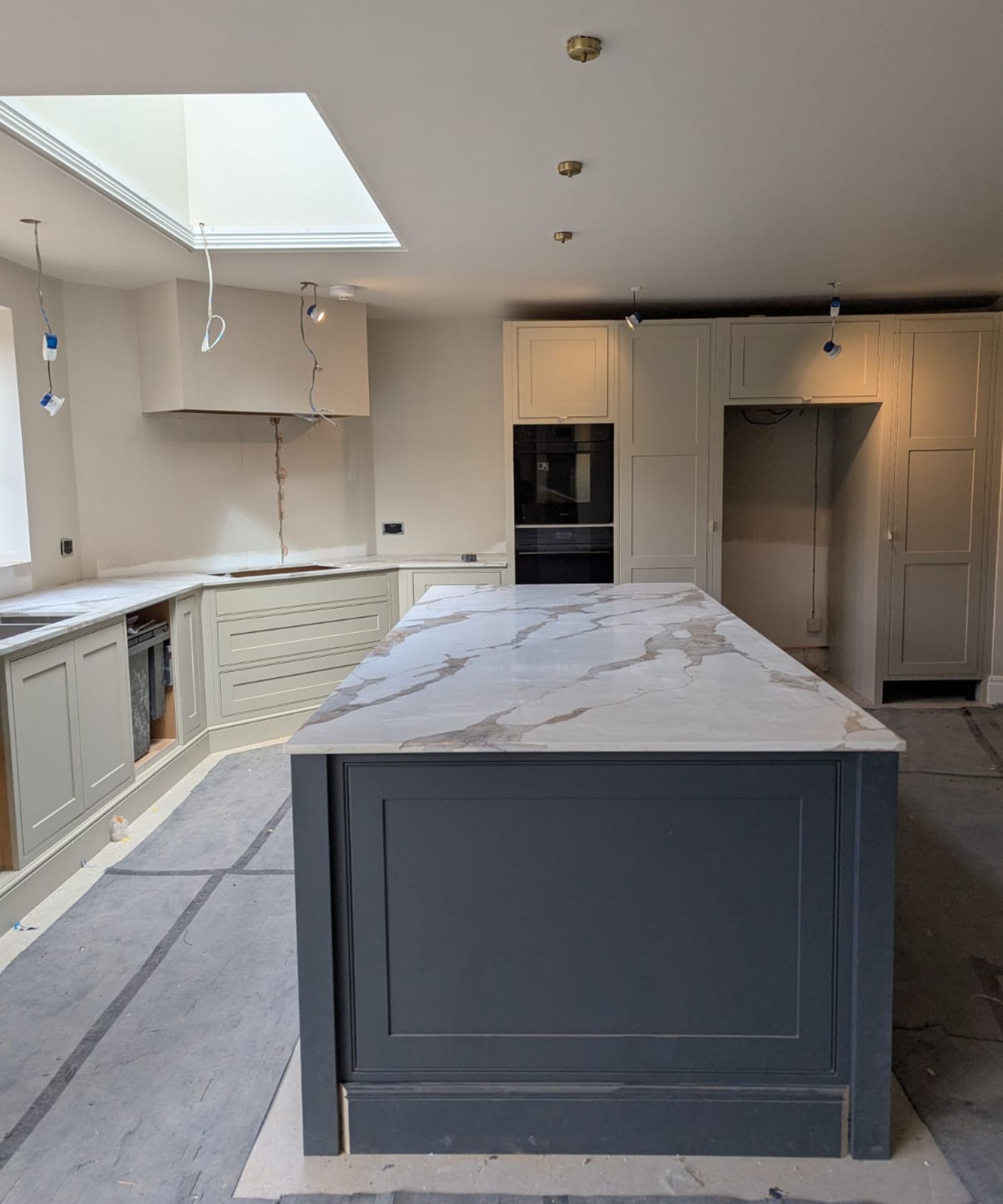 A large kitchen mid-fit with a dark grey island topped with marble-effect stone, sage green shaker cabinets, double ovens, a rooflight above and electrics still being finished.