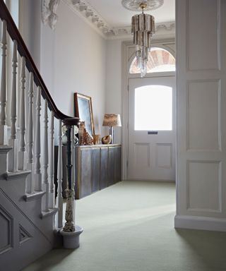 Large hallway with traditional architectural features, white walls with wall paneling, glass chandelier, carpeted floor, metallic console table decorated with ornaments and a table lamp, white painted staircase with dark wood handle