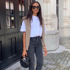 British female influencer Lorna Humphrey poses on the sidewalk in London wearing oversize black sunglasses, an oversize white T-shirt, mini black bag, tan sweater draped over the shoulders, and gray jeans