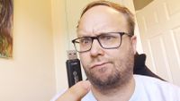 A confused white man with dark-rimmed glasses holding a Kingston USB drive in his hand in front of his face, looking at it.