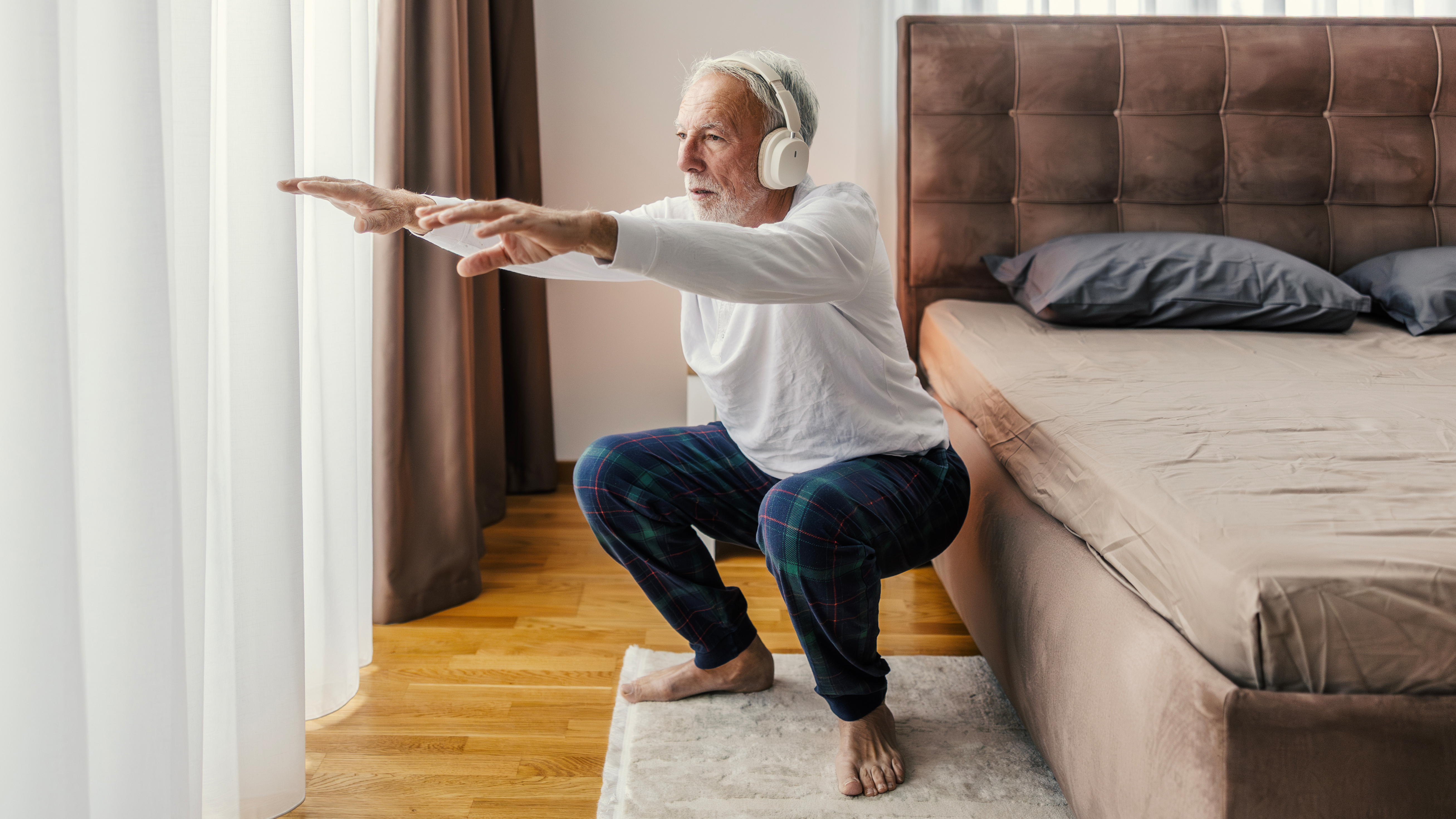 A man wearing pyjamas and overear headphones performs a squat next to his bed.