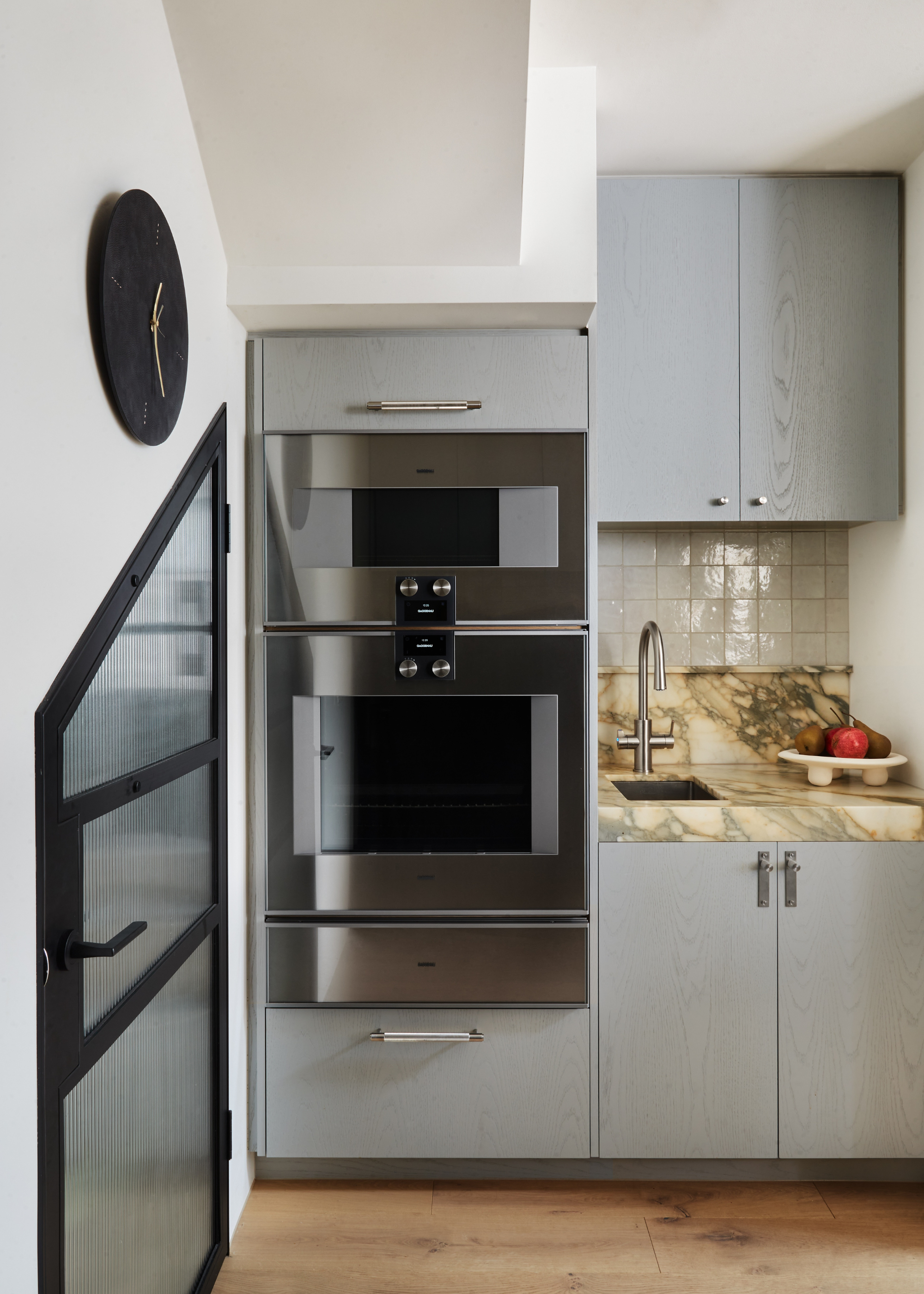 a light grey blue kitchen with a statement countertop next to a a double oven housing unit with a clock to the left and angled crittall style door with fluted glass