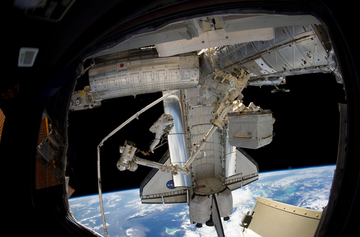 Discovery STS-133 astronaut Steve Bowen stands at the tip of the International Space Station&#039;s Canadian robotic arm, with the Earth and space shuttle in the background, during the second spacewalk of his mission on March 2, 2011. 