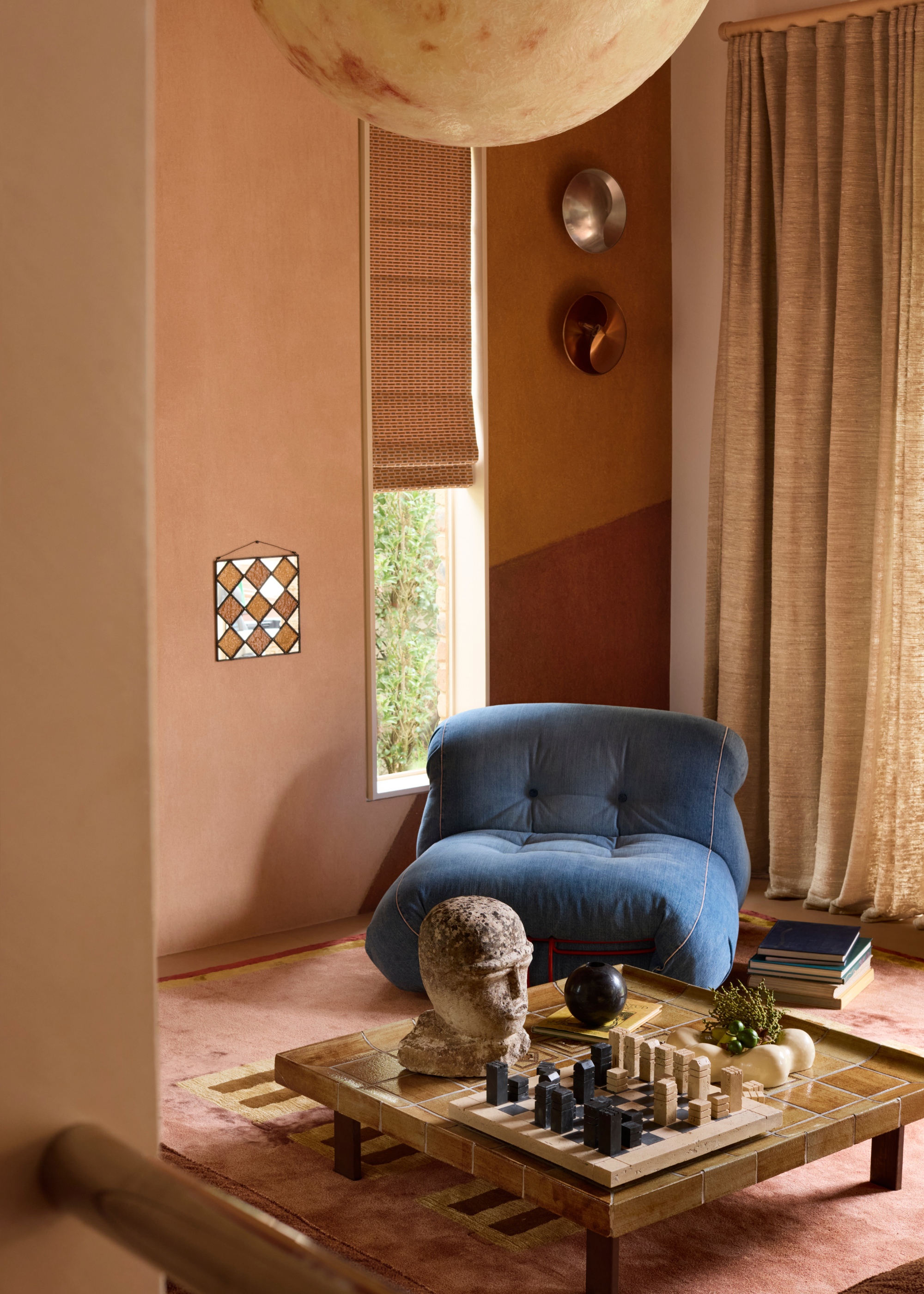 A living room corner in earthy tones with floor-length beige curtains, a narrow window with a roman blind half closed, and a large, plumped and low-profile blue lounge chair with a pile of books next to it, with a low-profile coffee table in front with a chess set