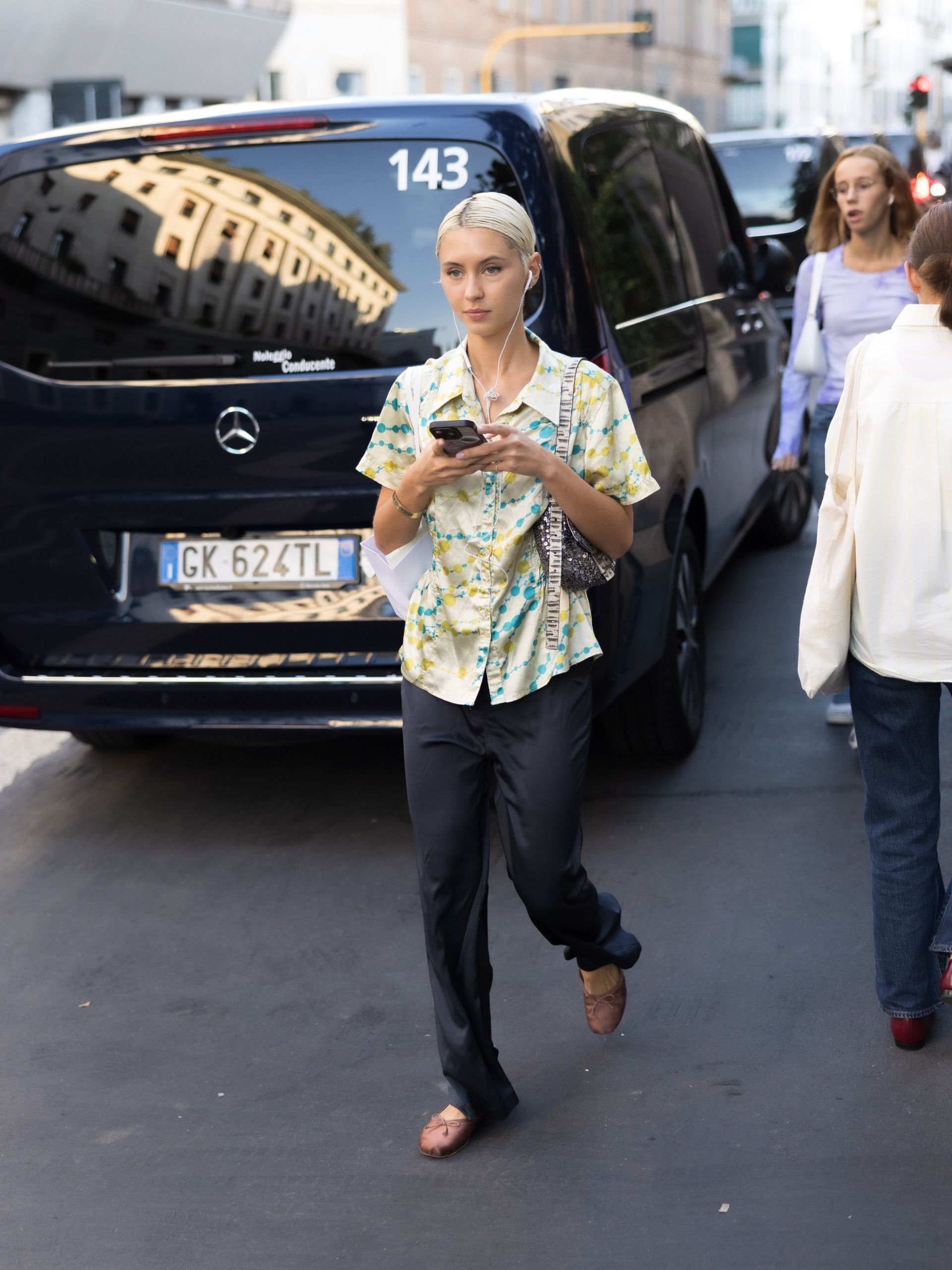 Iris Law steps outside in Milan wearing a short-sleeved satin button-down shirt with black satin trousers and brown satin ballet flats.