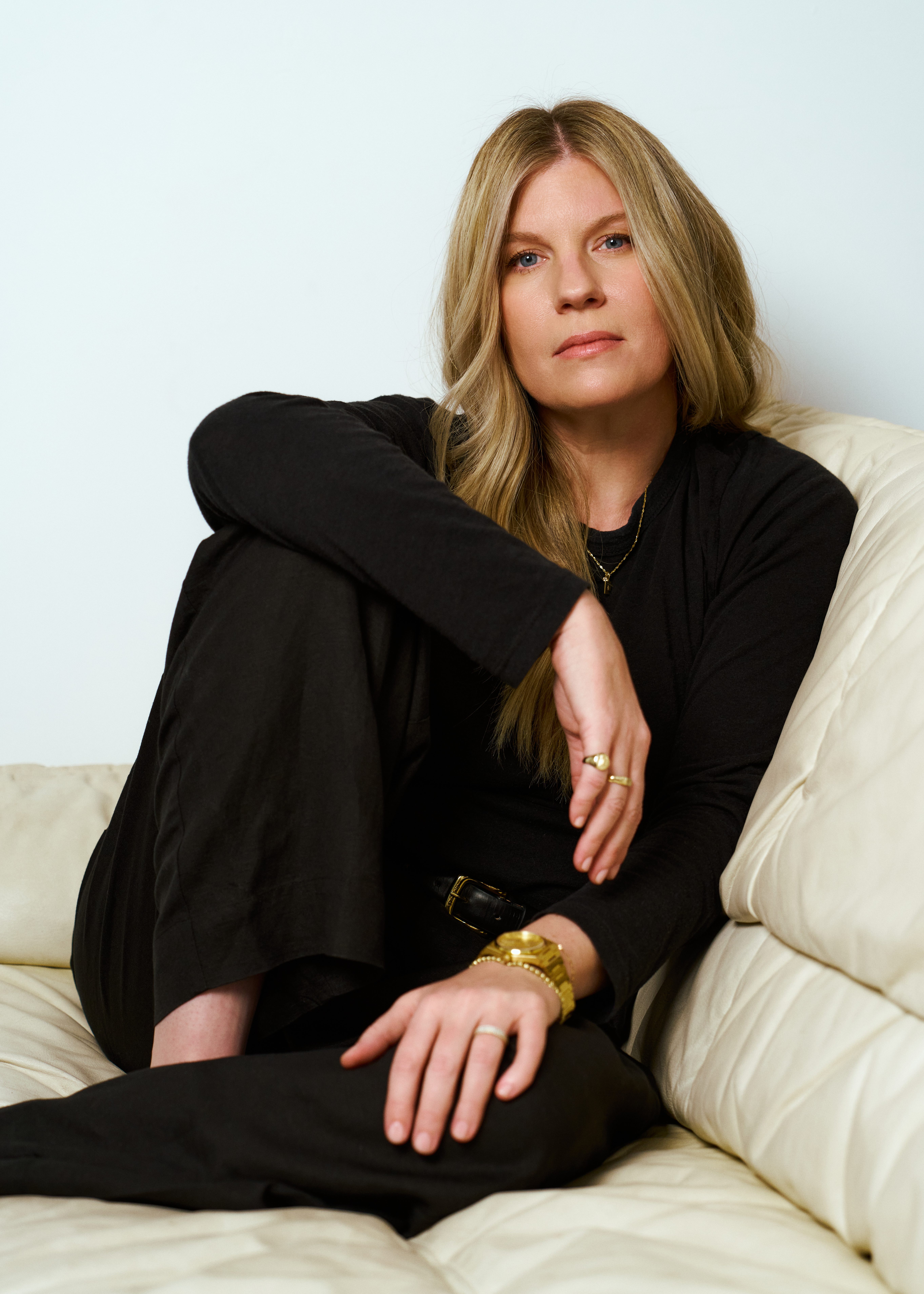 showrunner cassie pappas poses on a white leather couch wearing an all black outfit with gold jewelry