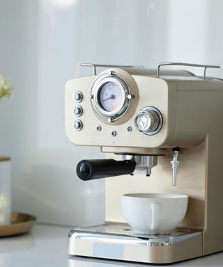 A cream coffee maker with a white curved cup underneath it, on a white counter with a white wall behind it