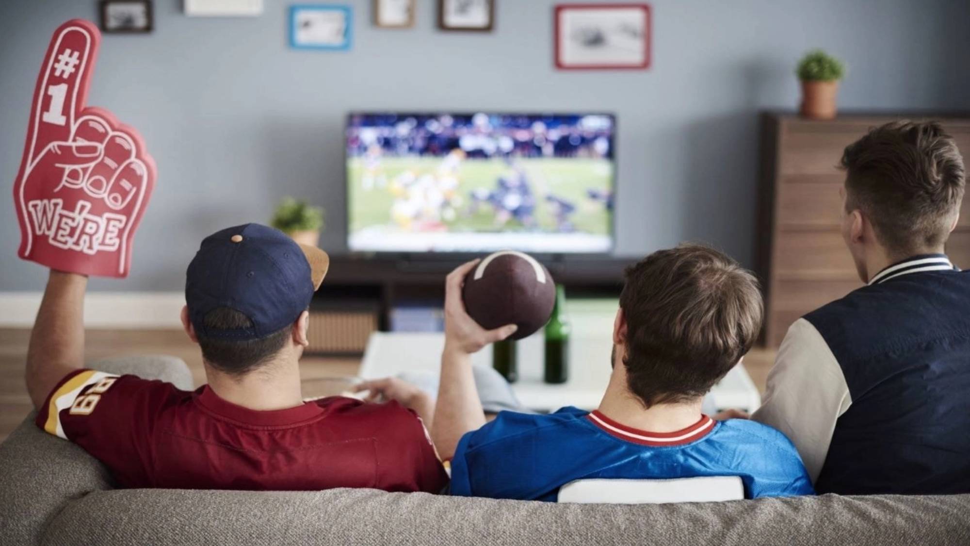 Three people watching football on TV, one has a foam finger, and one is holding a football