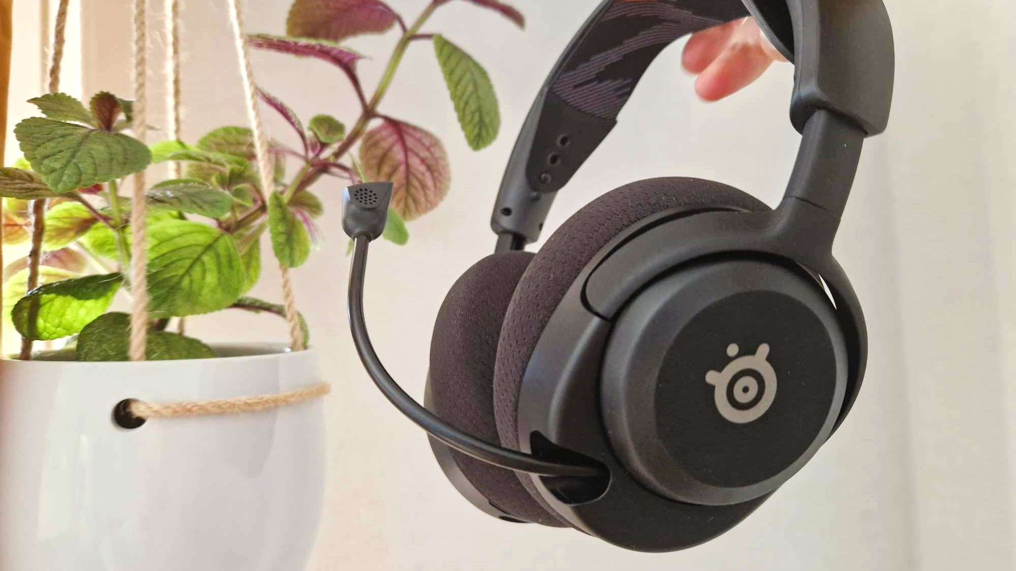 En hand håller upp ett par SteelSeries Arctis Nova 4 framför en hängande krukväxt vid en vit vägg.