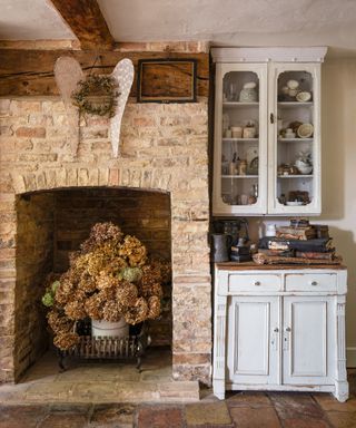 brick fireplace with display of dried hydrangeas in recess and beside a vintage dresser and glass fronted wall unit